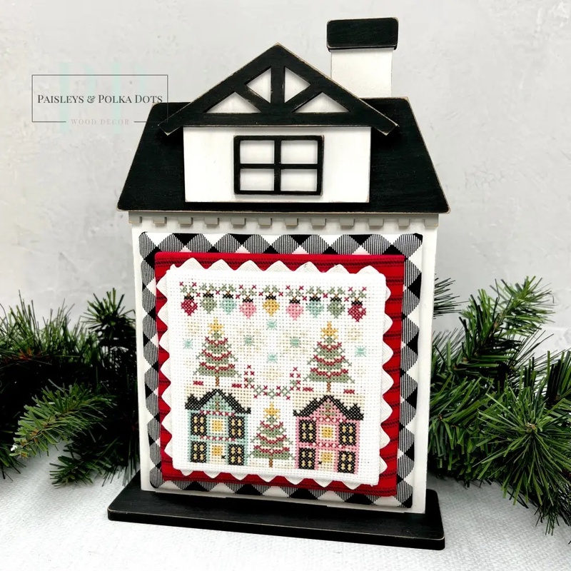 The fully assembled and painted City House #4 Cross Stitch Display Backer, staged on a white tabletop with a decorative garland, displaying a festive cross-stitch project.