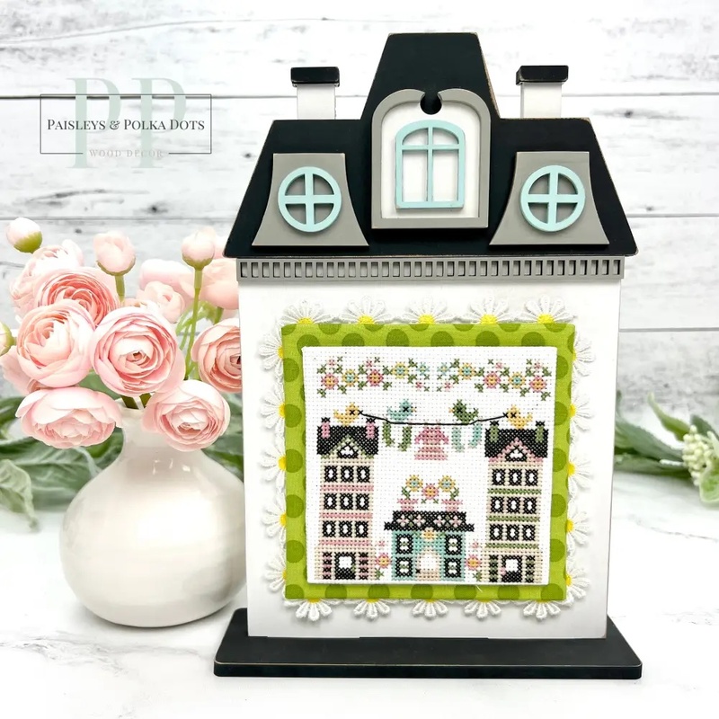 The fully assembled and painted Victorian City House Cross Stitch Display Backer, staged on a marble countertop with coordinating flowers, displaying a spring cross-stitch project.