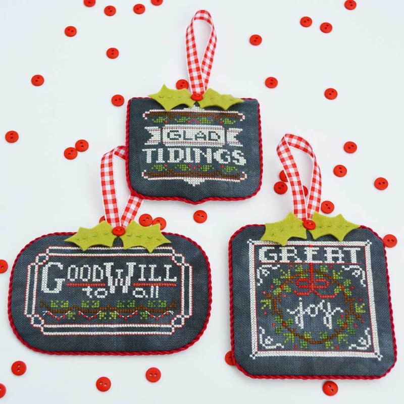 Three cross stitch decorative holiday ornaments with messages: Goodwill to All, Glad Tidings, and Great Joy, surrounded by red sequins.