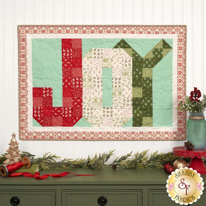 The completed Patchwork Holiday Joy project colored in red, cream, green, and aqua fabrics from the Sugarberry collection by Andover Fabrics, hung on a white paneled wall and staged with coordinating Christmas decor and foliage.