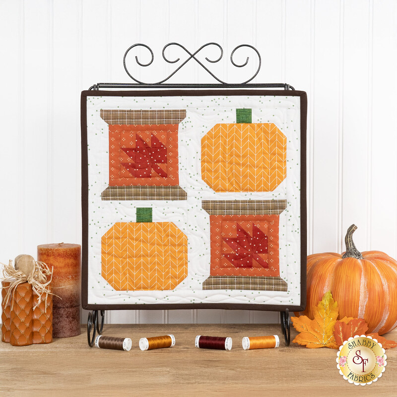 The completed Sew Thankful wall hanging, two spools with pieced leaves paired with two pieced pumpkins in bold autumn colors, hung on a display stand and staged with coordinating spools of thread and autumn decor.