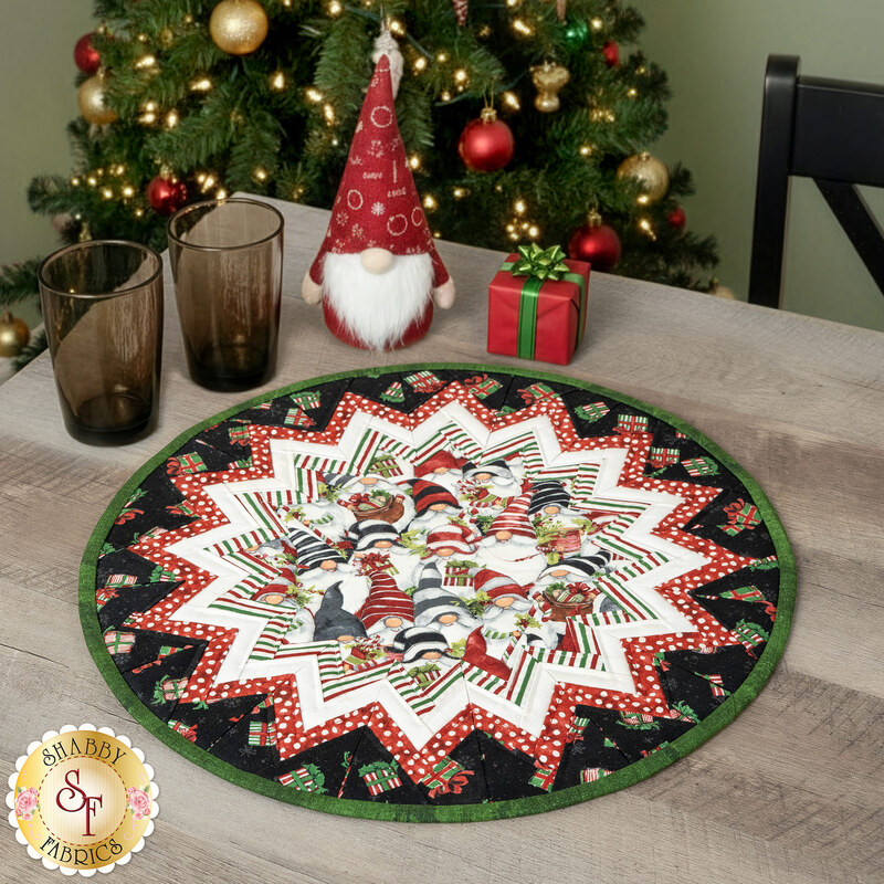 The completed Point of View Folded Star Table Topper colored in white, red, black, and green fabrics from the Merry Gnome-ments collection by Wilmington Prints, staged on a gray wood table with coordinating kitchen decor.