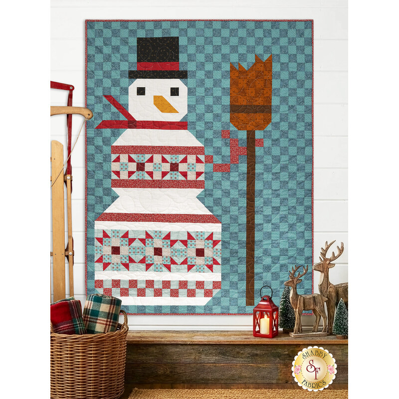The completed Patchwork Snowman quilt colored in the cool winter teal, red, and white of the Frosty and Forest Friends collection, hung on a white shiplapped wall and framed by coordinating winter décor.