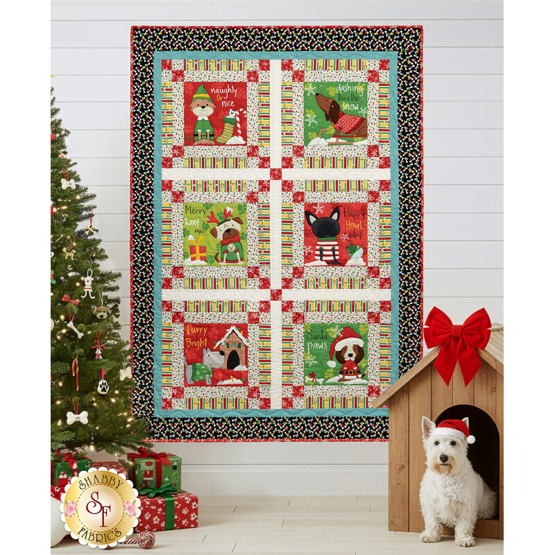 The completed Merry Woof-Mas Panel quilt, colored in bold red, green, yellow, blue, black, and white fabrics from the Merry Woof-Mas collection by Studio E Fabrics, hung on a white paneled wall and staged with a Christmas tree with dog-themed ornaments. A doghouse with a red bow and a Westie wearing a Santa hat sit in the lower right corner.