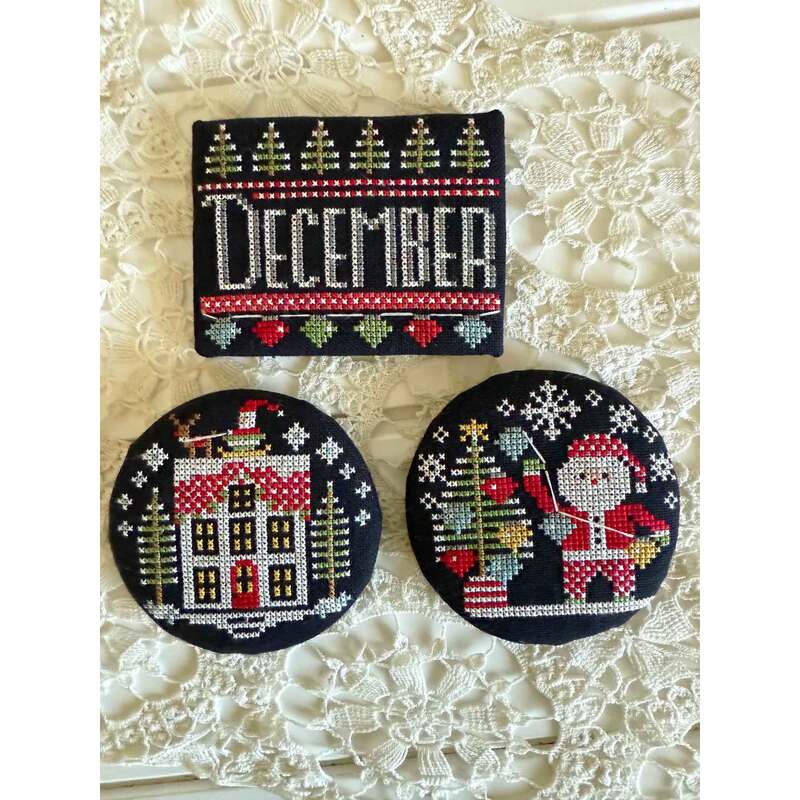 Three decorative cross stitched Christmas-themed items: a sign reading December and two circular images featuring a house and Santa.