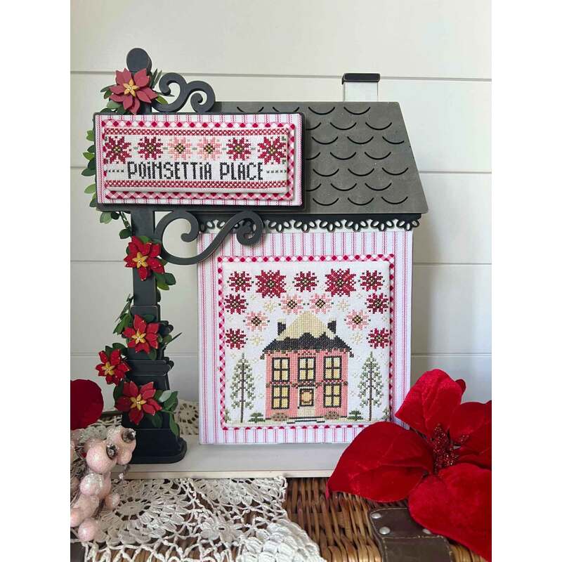 A cross stitch pattern featuring a decorative sign that reads Poinsettia Place, featuring a quaint house surrounded by flowers.