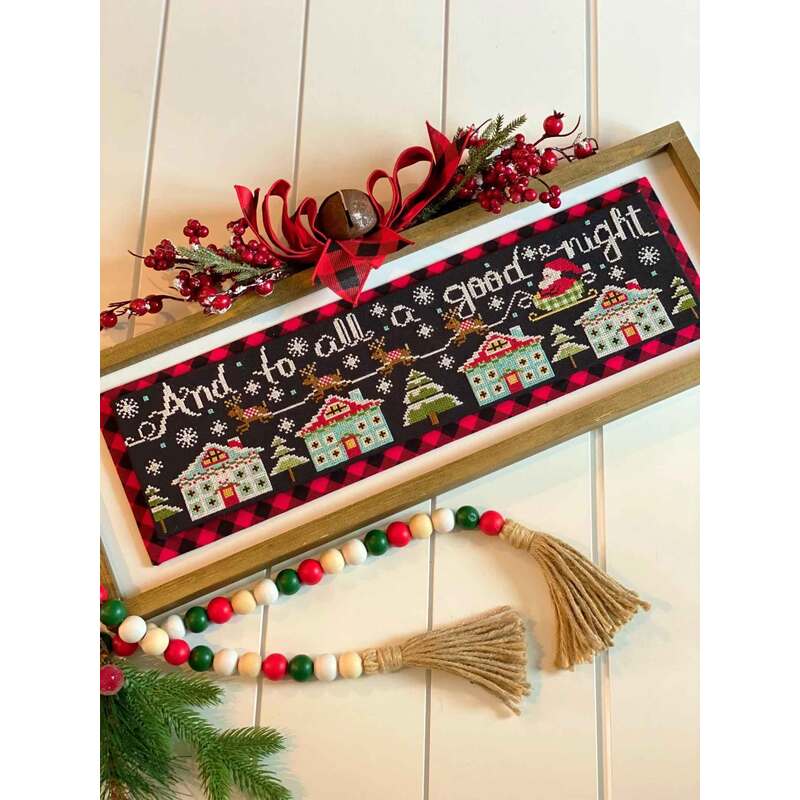 A decorative cross stitch sign with And to all a good night, surrounded by winter-themed houses and greenery.