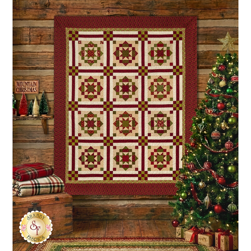 The completed Christmas in the Mountains quilt, colored in red, cream, and green fabrics from the Baubles & Balsam collection from Henry Glass. The project is staged on a cabin wall with a coordinating Christmas tree and décor.
