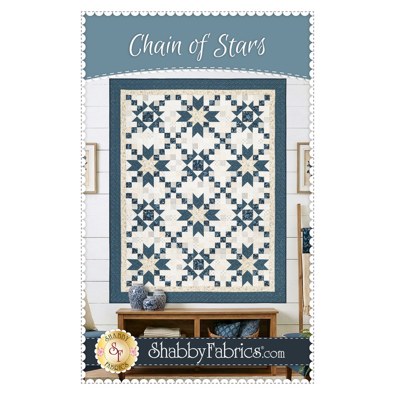 Front cover of the Chain of Stars pattern, showing the completed quilt colored in blue fabrics from the Sacre Bleu collection and cream fabrics from the Itty Bitty Backgrounds collection, staged on a white shiplap wall and staged with coordinating furniture and décor.