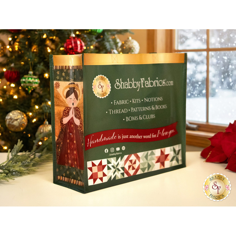 The front side of the Shabby Fabrics Merry Christmas Tote Bag, a green bag featuring gold trim, green and red patchwork blocks, and info about the website, staged on an off-white table in a warmly lit room with a decorated Christmas tree and a window with a winter wonderland outside.