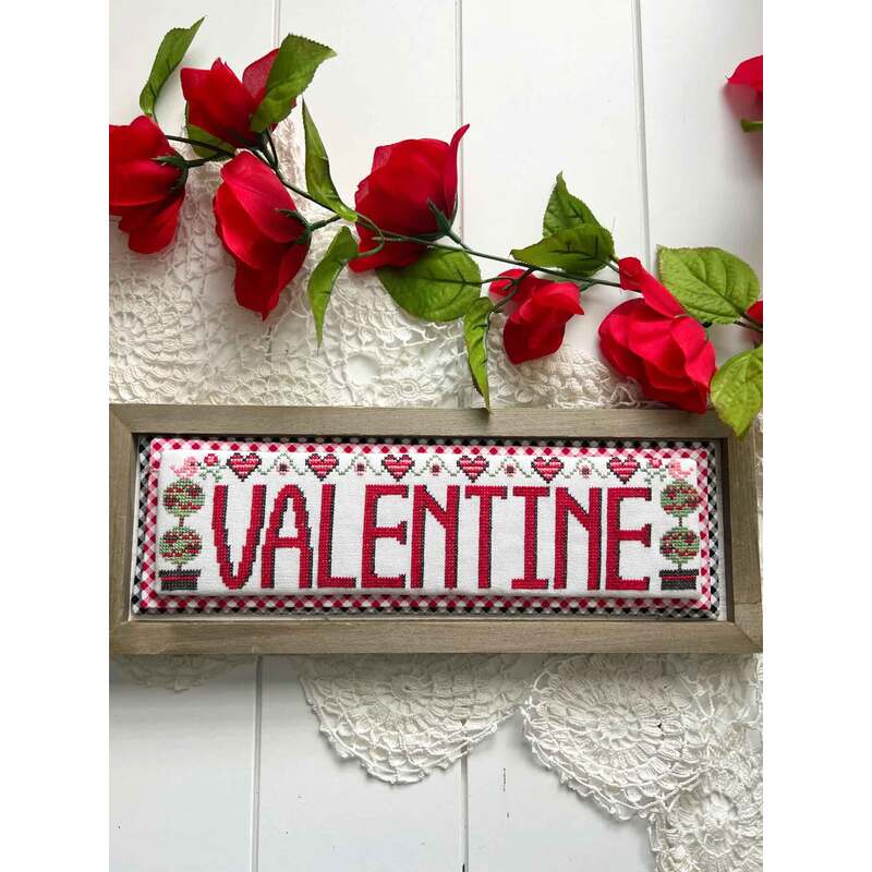 The front of the Valentine Word Swap cross stitch pattern showing the completed project surrounded by coordinating holiday décor. 