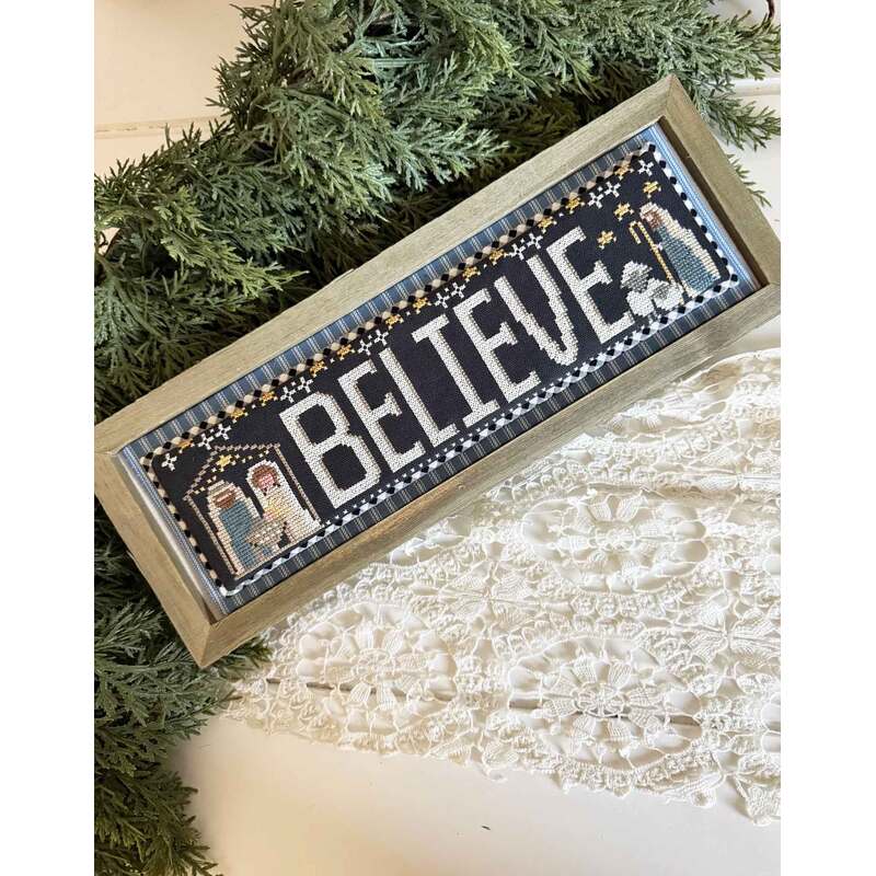 A framed cross stitch sign displaying the word BELIEVE with decorative elements and a lace background.