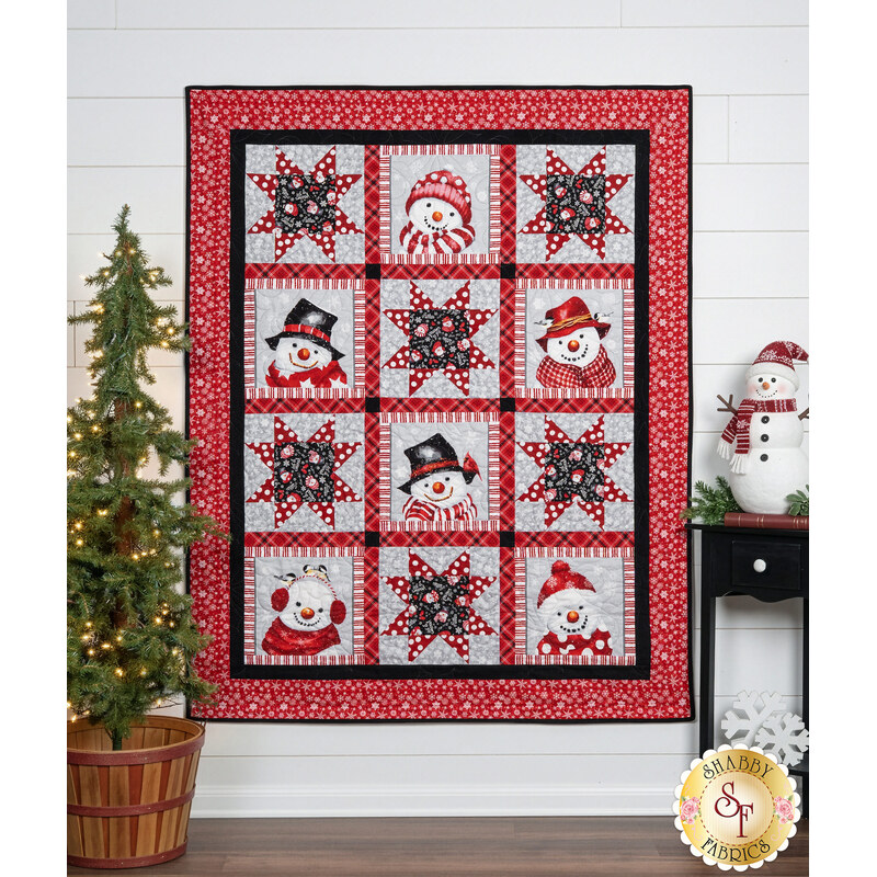 The completed Snow Crew Besties Quilt, colored in red, white, gray, and black fabrics from the Snow Crew Flannel collection, hung on a white paneled wall and staged with coordinating decor like a decorated tree, and a snowman figurine.