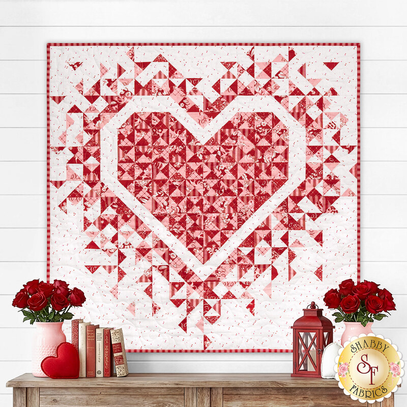 The completed Mini Exploding Heart quilt colored in red, pink, and white fabrics from the Letters to Cupid collection, hung on a white paneled wall above a wooden dresser; coordinated pink and red décor adorn the table. 
