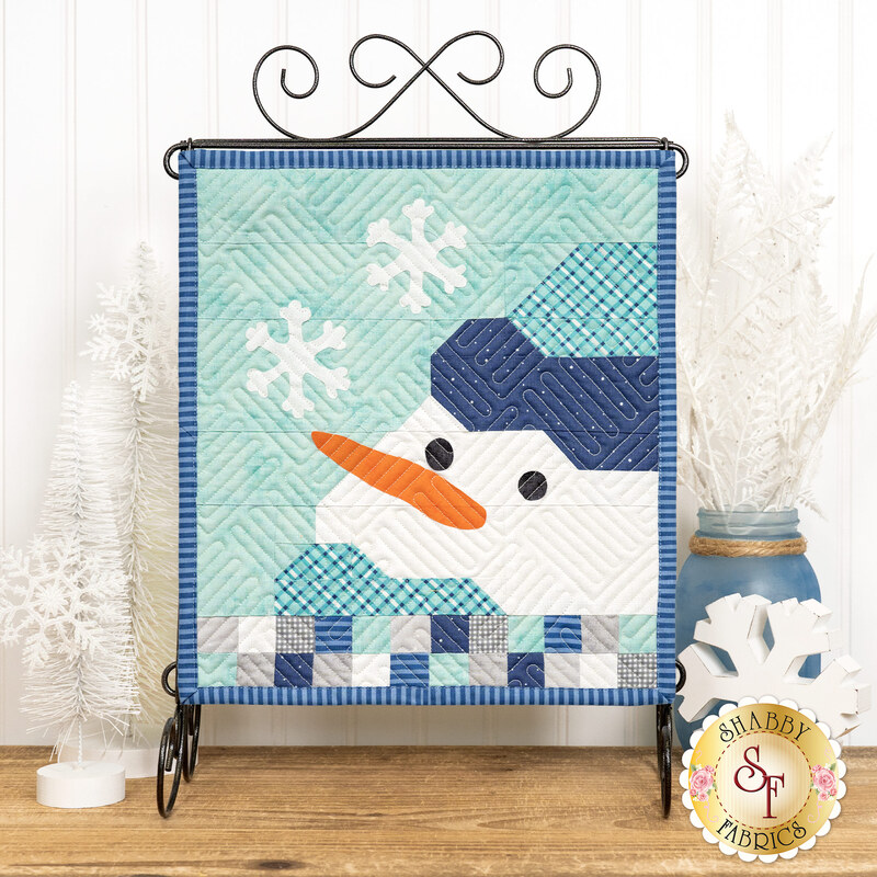Winter themed table top banner featuring an appliqué snowman and snowflakes in cool blue and gray colors, hung on a display hanger and staged with coordinating decor.