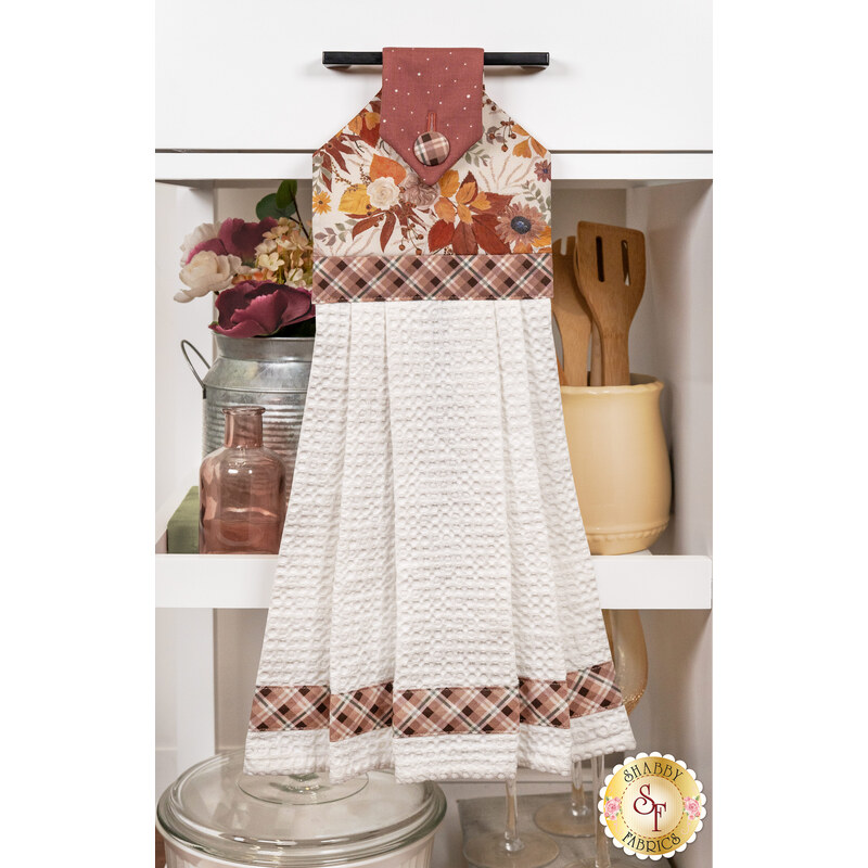 The Hanging Towel, colored in brown, burnt orange and white fabrics from the Evening On The Prairie collection, staged on a white kitchen drawer with coordinating decor.