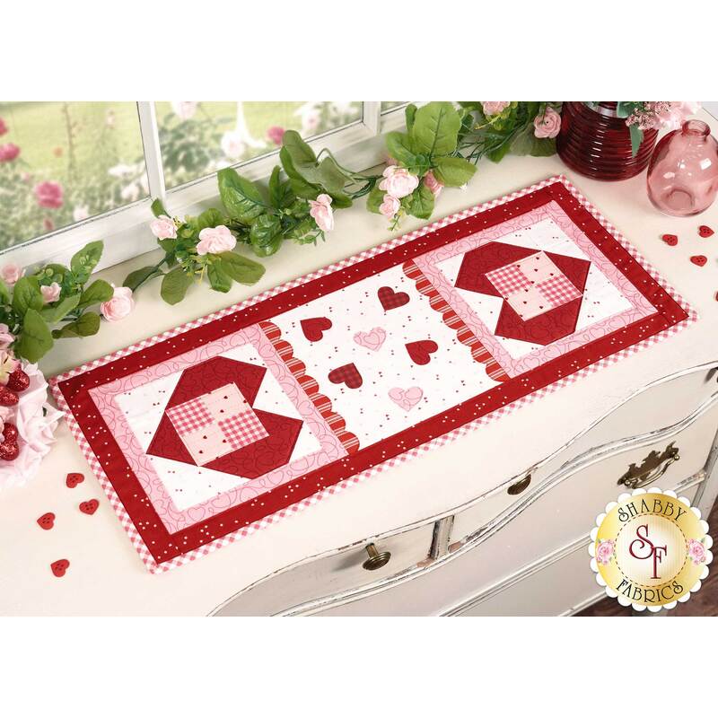 The completed Sew Cute Runner - February table topper, staged on a white dresser, accented with scattered red buttons, a strand of greenery with pink flowers, and two vases sitting in the upper right corner.