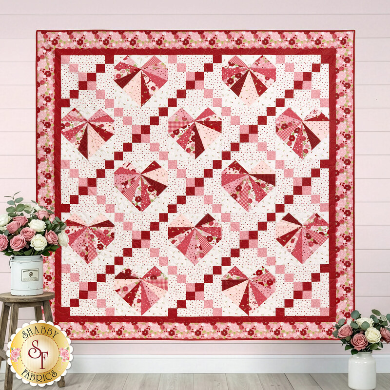 The completed Foundation Paper Piecing Heart & Lattice Quilt colored in pink, red, and white fabrics from the Sew In Love collection by Riley Blake Designs, hung on a pink shiplap wall and staged with coordinating white buckets of pink and white roses.