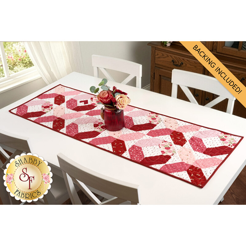 The completed Hugs & Kisses Table Runner, colored in pink, red, and white fabrics from the Sew In Love collection by Riley Blake Designs, staged on a white table beside a bright window. A yellow banner in the corner reads "Backing Included!"
