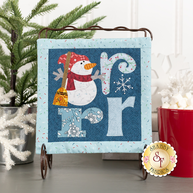 The On Wander Lane Mini Quilt - Frost Hill - Brrr Kit featuring a cheerful snowman in a scarf next to colorful letters and snowflakes.