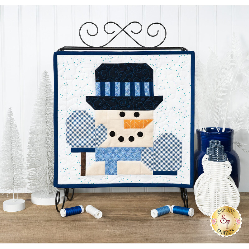The completed January project featuring a snowman dressed in a hat, gloves, and scarf, hung on a craft holder and staged on a wooden table with coordinating spools and holiday décor.