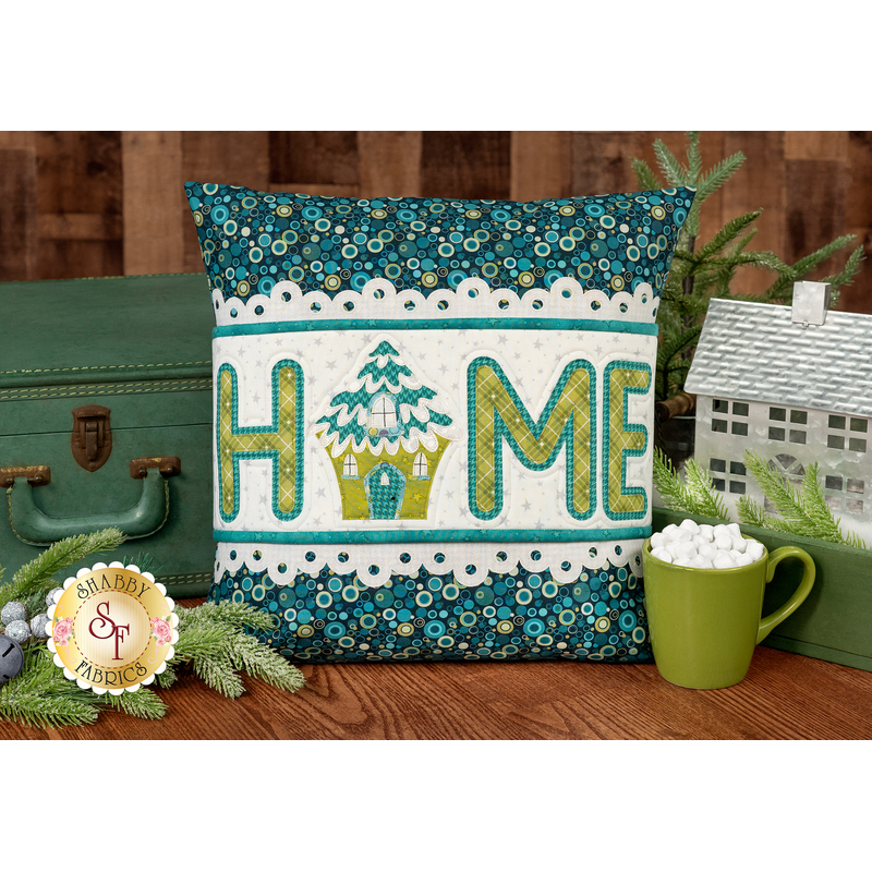 The completed Home Pillow Wrap & Cover, colored in white, teal, and green fabrics. The word "Home" is spelled out in appliqué letters and the "o" is replaced with a tree house. The project is staged with a vintage suitcase and winter themed decor.