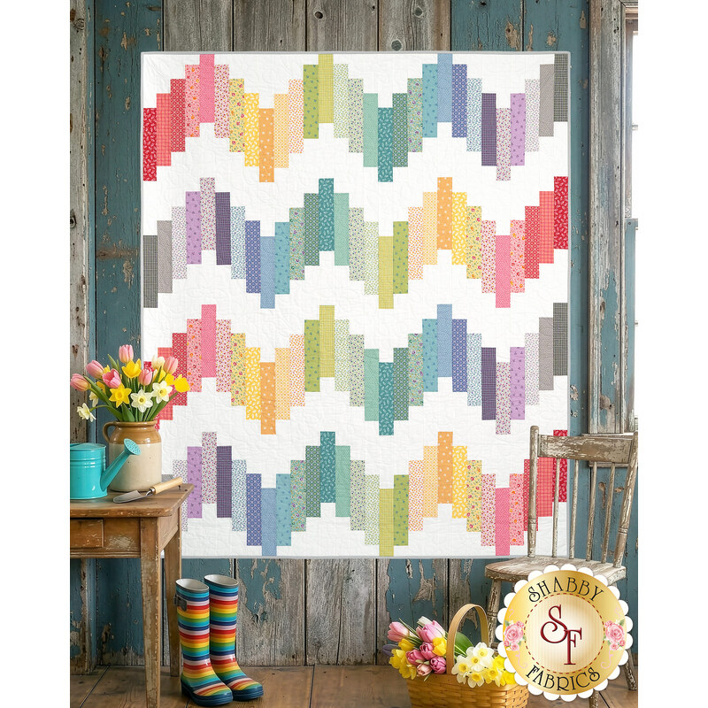 The completed Ridiculously Easy Jelly Roll Quilt in fabric from the Sweet Things collection, staged on a rustic paneled wall with wooden chair, cut flowers, and rainbow rubber boots. 