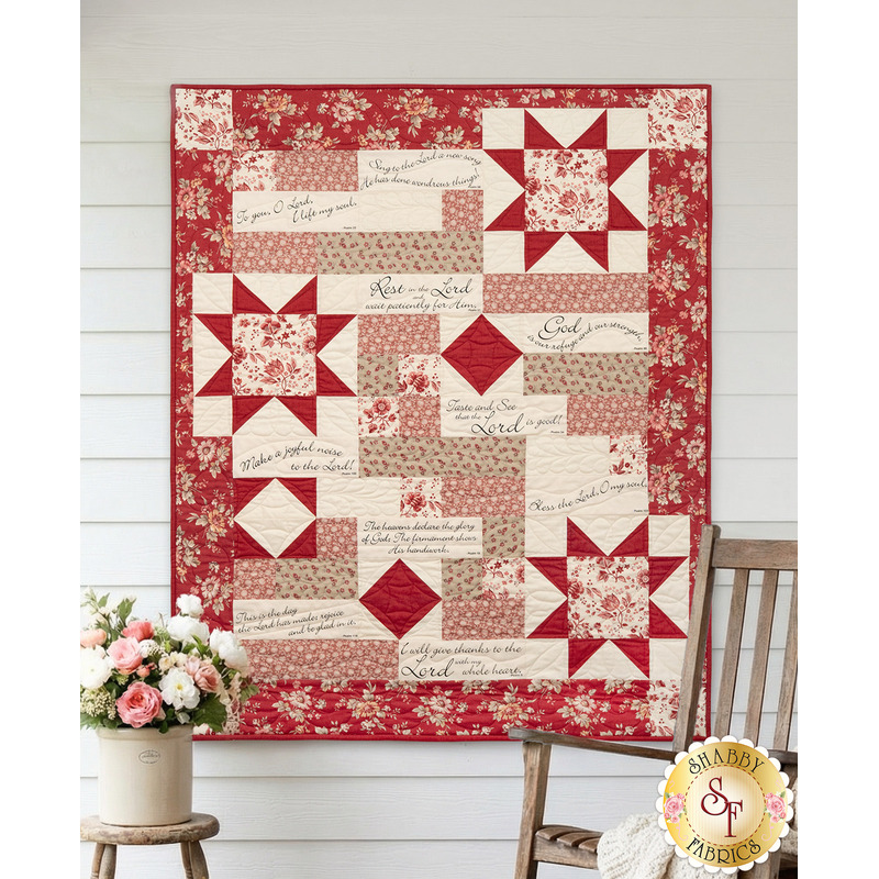 The completed Comfort of Psalms Quilt, colored in red, pink, and cream fabrics from the Joie de Vivre collection. The quilt is displayed on a white paneled wall and staged with coordinating pink flowers and a rocking chair.