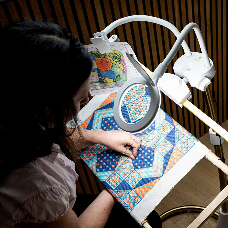 A person stitches a colorful quilt pattern under a magnifying lamp in a well-lit workspace.