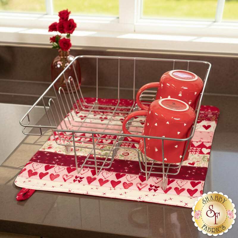 The completed Dry Ideas mat colored in bold pink, red, and cream fabrics from the Darling Peony collection, staged on a kitchen countertop beside a sink with a dish rack on top holding coordinating Valentine's Day themed mugs.
