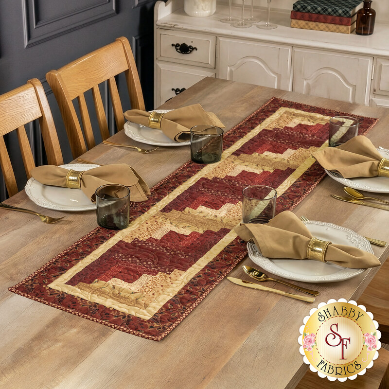 The completed Log Cabin Table Runner, colored in rich red and deep tan fabrics from the Dear Scarlet collection by Marcus Fabrics, staged on a wooden table with four coordinating table settings.