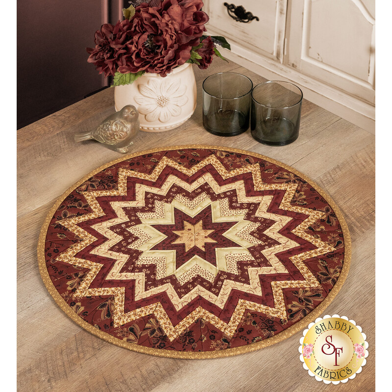 The completed Folded Star Table Topper colored in rich red and deep tan fabrics from the Dear Scarlet collection by Marcus Fabrics, staged on a wooden table with coordinating glassware and décor.