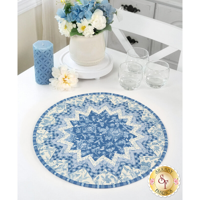 The completed Point of View Folded Star Table Topper, colored in blue and white fabrics from the Penelope collection by Riley Blake Designs, staged on a white table beside coordinating glassware, flowers, and a candle.