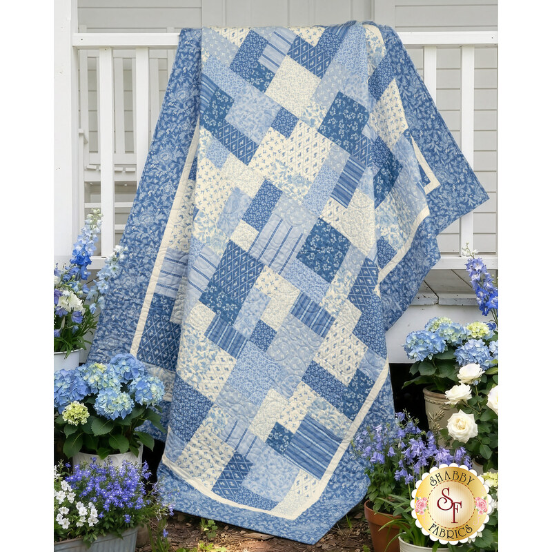 The completed Easy as ABC and 123 Quilt, colored in blue and white fabrics from the Penelope collection by Riley Blake Designs, draped from a white porch fence and surrounded by coordinating florals.