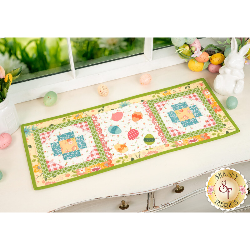 The completed Sew Cute Runner - April table topper, staged on a white dresser, accented with scattered colored eggs, a wreath of easter eggs and a ceramic bunny statue sit in the top right corner. 