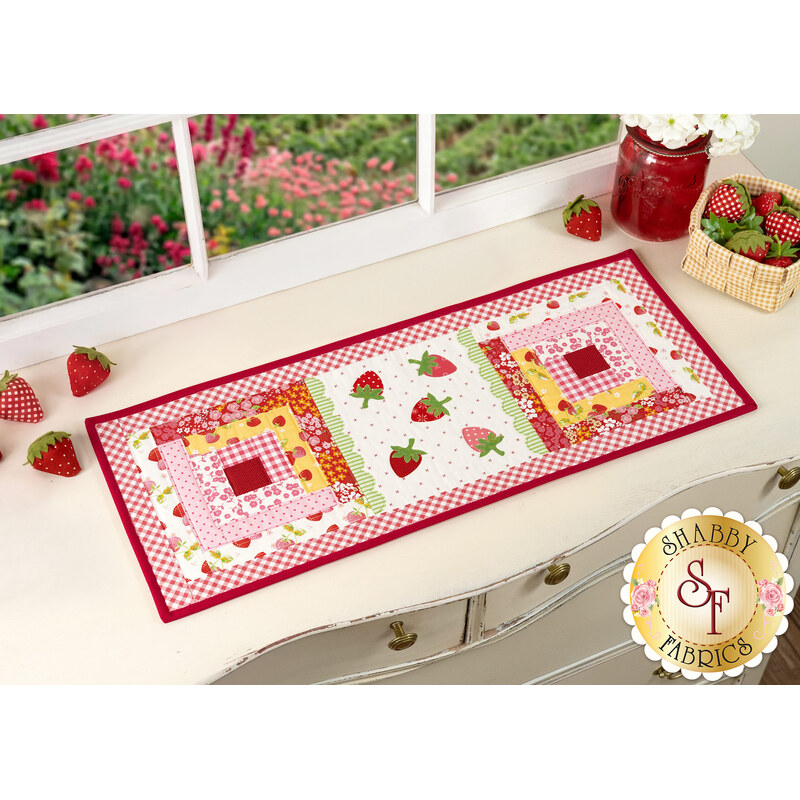 The completed Sew Cute Runner - June table topper, staged on a white dresser, accented with scattered red strawberries, and a red vase sitting in the upper right corner.