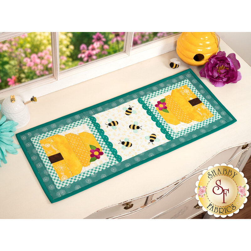 The completed Sew Cute Runner - August table topper, staged on a white dresser, accented with purple and blue flowers and a ceramic bee hive statue sit in the top right corner. 