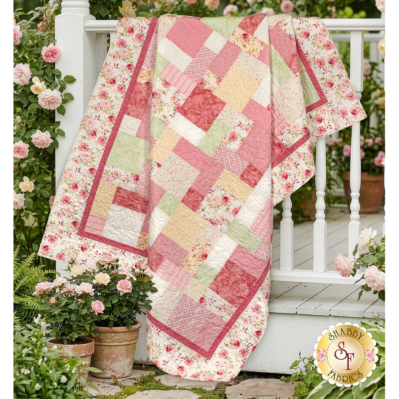 The completed Easy as ABC and 123 quilt kit colored in soft red, pink and white fabric from the  Yours Truly collection by P&B Textiles, draped over a white porch surrounded by pink roses.