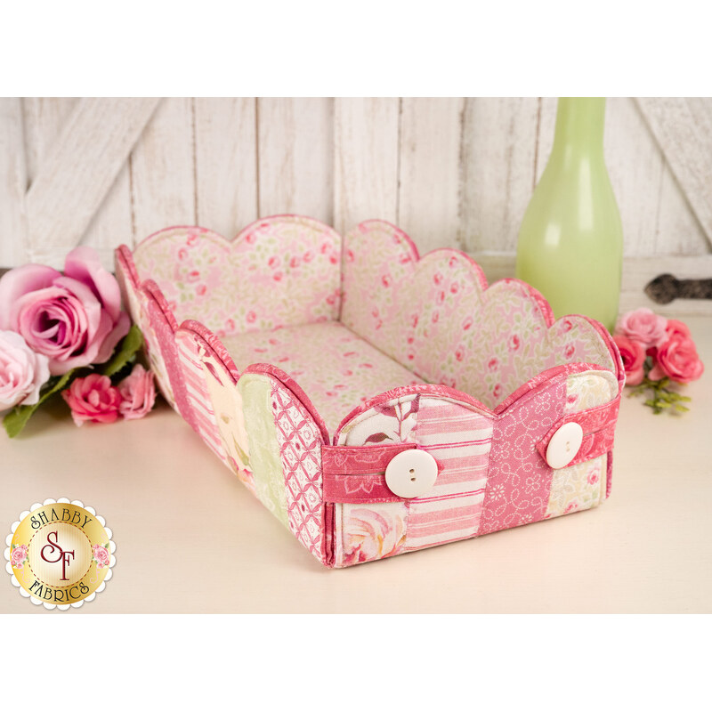 The completed Scalloped Basket, colored in pink and white fabrics, staged on a white table with coordinating flowers and décor.