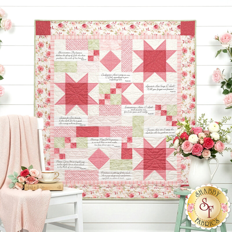 The completed Prayers of the Heart Quilt, colored in pink, light pink, and white fabrics from the Yours Truly collection. The quilt is displayed on a white paneled wall and staged with coordinating flowers, a pink knit blanket is draped over a chair.