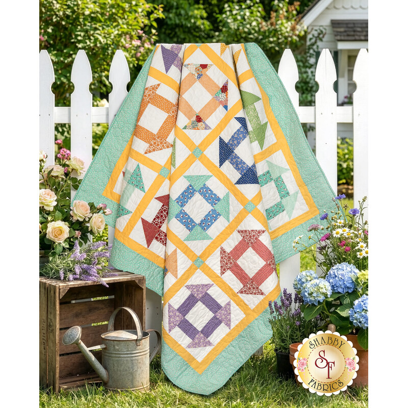The completed Churn Dash Layer Cake Quilt colored in vibrant prints from the Aunt Grace Goes Scrappy collection by Marcus Fabrics, draped over a white picket fence and surrounded by coordinating bunches of flowers.