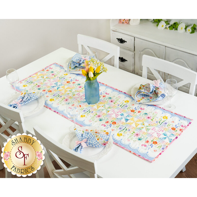 A Spring themed table runner featuring rainbow watercolor fabrics and patchwork pinwheel stripe blocks from the Spring Cheer collection by Clothworks, staged on a white table set with coordinating napkins, plates, glasses, and a vase of tulips. 