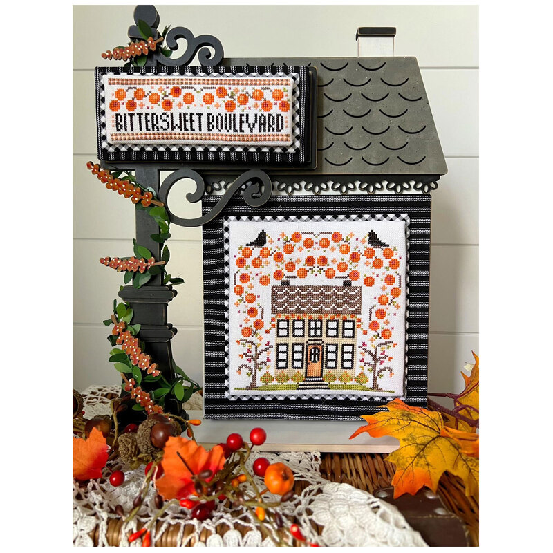 A framed cross stitch art piece featuring a fall decorated house next to a similarly dressed street post labeled BITTERSWEET BOULEVARD.