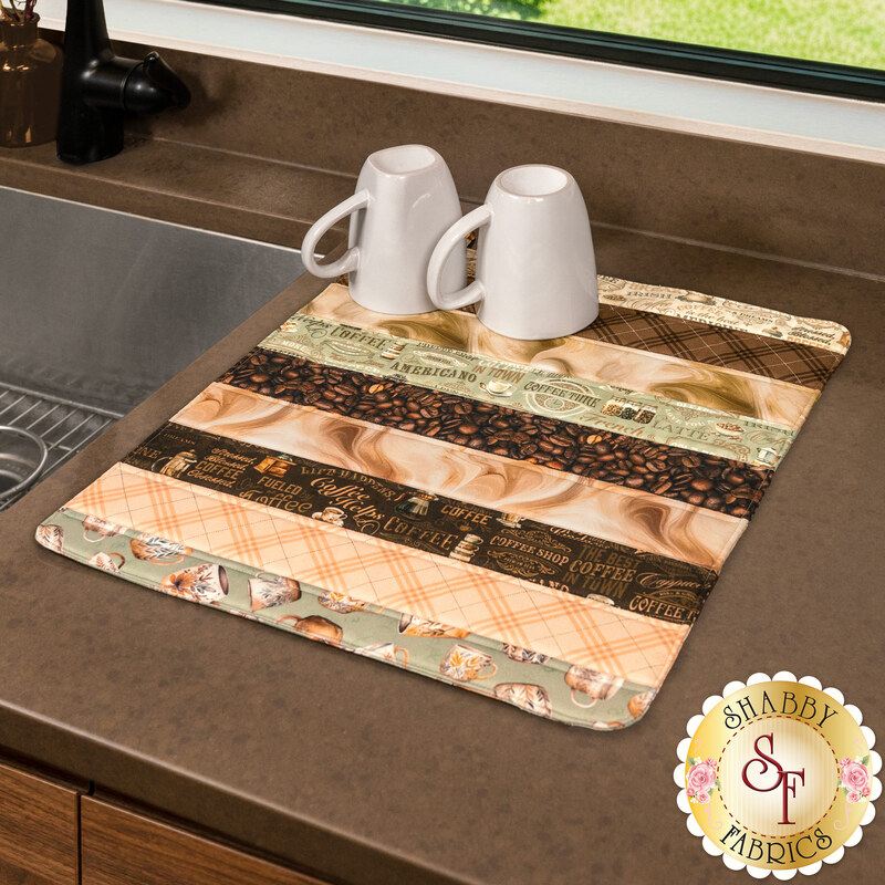 The finished Dry Ideas mat, made with brown, tan, and sage green coffee-themed fabrics from the Coffee Time collection from Riley Blake Designs, staged on a brown countertop with two ceramic mugs. A gold shabby fabrics logo is in the corner. 