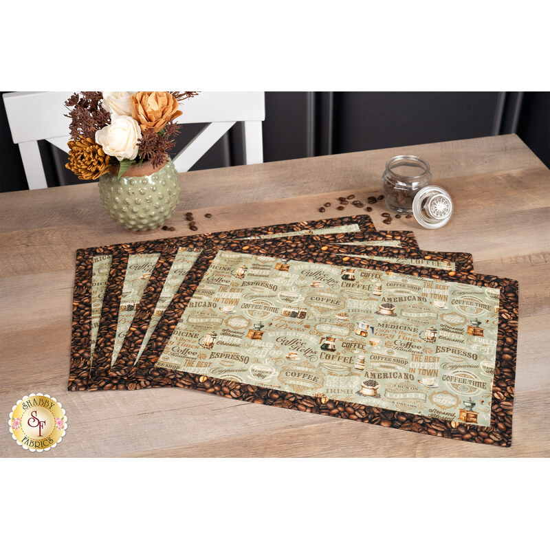 A shot of the four completed overlapping placemats made with coffee-themed taupe fabric with printed text and coffee bean fabric borders, staged on a wooden table with coordinating décor.