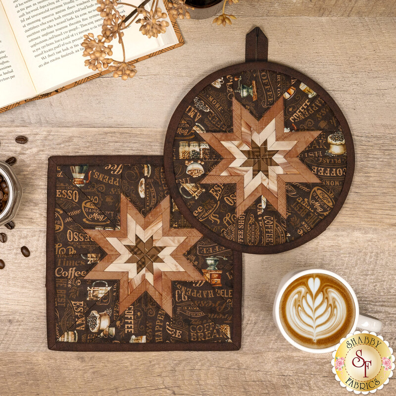 The finished Square and Round Folded Star Hot Pads made with the Coffee Time collection in brown, featuring chocolate brown and cream stars at the center of the hot pads surrounded by dark brown fabric with coffee related phrases. The hot pads are staged on a wooden table with coordinating décor, a gold shabby fabrics logo is in the bottom right corner.
