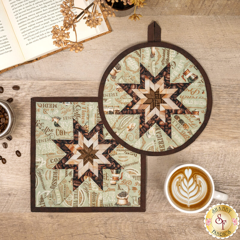 The finished Square and Round Folded Star Hot Pads made with the Coffee Time collection in sage, featuring chocolate brown and cream stars at the center of the hot pads surrounded by sage green fabric with coffee related phrases. The hot pads are staged on a wooden table with coordinating décor, a gold shabby fabrics logo is in the bottom right corner.