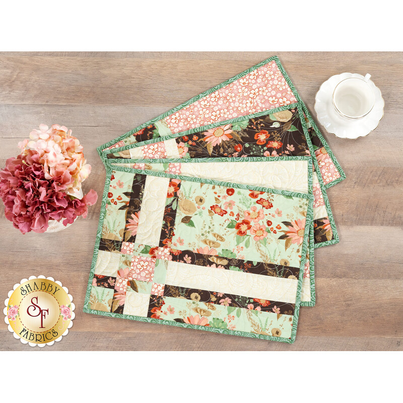 The four completed Fat Quarter Shuffle Mats, colored in cottage green, espresso brown, and blush pink fabrics from the Morning Latte collection by Riley Blake Designs, fanned out on a wooden table with coordinating flowers and a teacup.