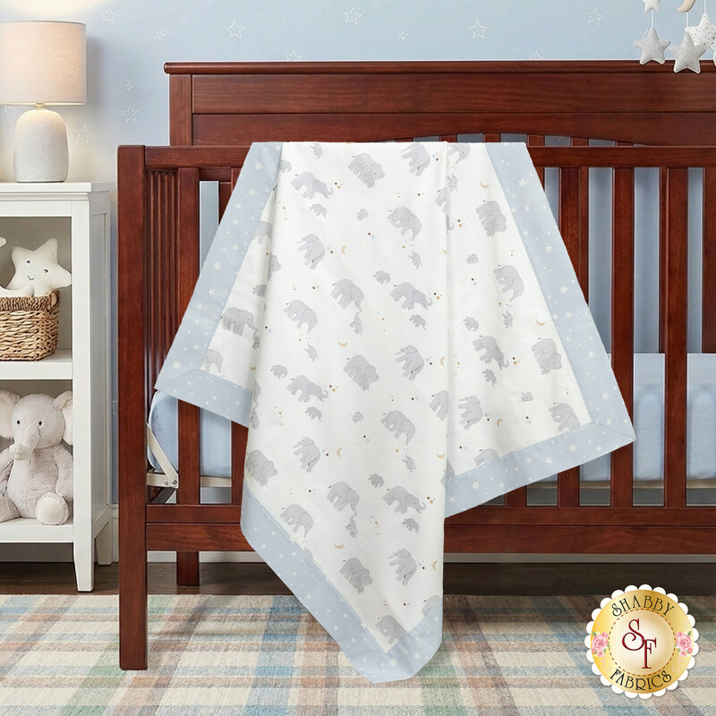 The completed self-binding receiving blanket in gentle pastel colors from the To The Moon And Back collection by Moda, artfully draped over the side of a wooden crib, staged with coordinating white furniture, fabric, and a stuffed elephant.