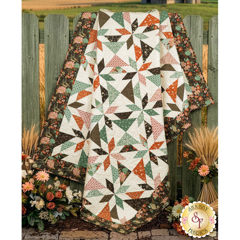 The finished Playing Favorites Quilt artfully draped over a wooden fence in a grassy field, surrounded by bundles of wheat with coordinating florals.